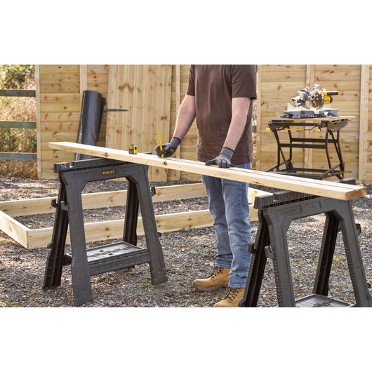 STANLEY 0-30-687 folding saw horse pair supporting wood board in outdoor workspace, person measuring board.