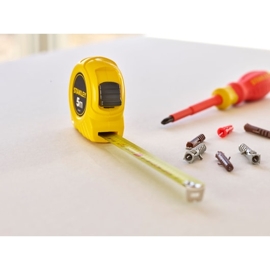 STANLEY 1-30-497 tape measure in yellow with extended blade, screwdriver, and fasteners on tabletop, oblique angle.