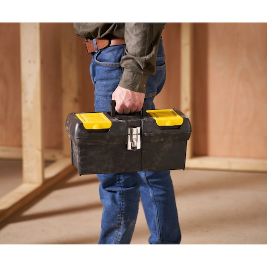 A person holding a black Stanley toolbox with yellow compartments, standing indoors in a construction or workshop setting.