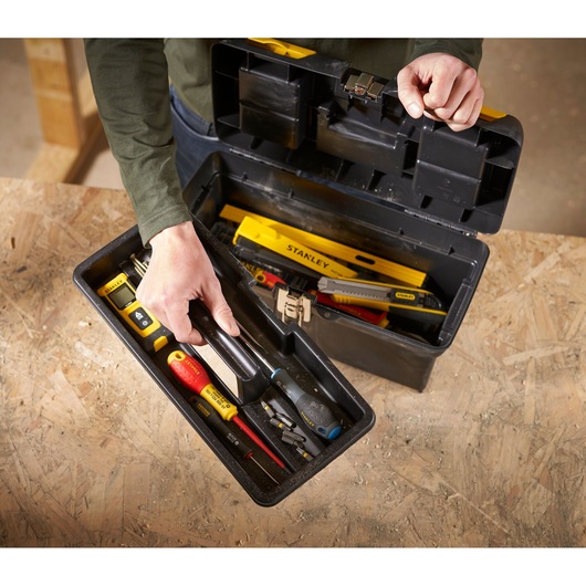 A person is opening a black Stanley toolbox on a wooden surface, revealing an assortment of hand tools including screwdrivers, a spirit level, and utility knives organized inside.