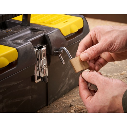 Close-up of hands locking a black and yellow STANLEY toolbox with a padlock and key on a workbench.