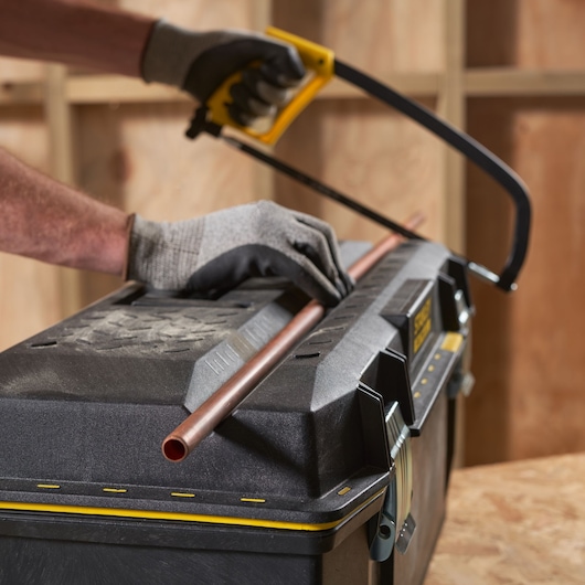 STANLEY 1-93-935 toolbox used as work surface, gloved hands cutting copper pipe with hacksaw, front angle