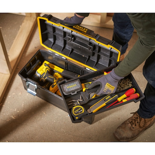 A person wearing gloves lifts the tray of a Stanley toolbox, revealing various hand tools, screws, a measuring tape, screwdriver, utility knife, and a power drill inside the box. The toolbox has a black and yellow design and is placed on a workshop floor.