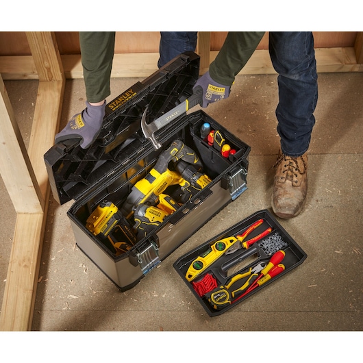 A person wearing gloves is opening a STANLEY toolbox filled with various tools, including a hammer, drill, screwdrivers, and other hand tools. The toolbox has removable trays and is being used in a construction environment.