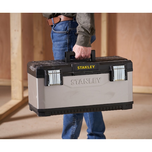 A person holding a STANLEY toolbox by its handle. The toolbox is grey with black accents and metal latches.