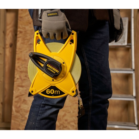 STANLEY 2-34-824 yellow 60m open reel tape measure held by worker in gloves, indoor workshop setting.
