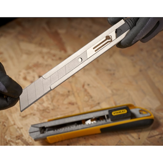 STANLEY 3-11-725 snap-off utility knife blade held above yellow and black utility knife on wood surface.