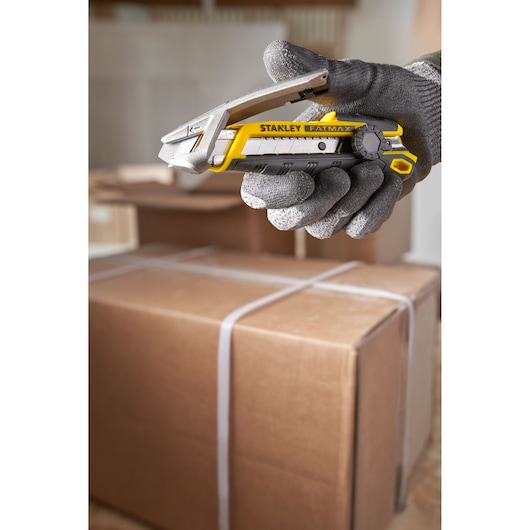 STANLEY FMHT10592-0 utility knife held in gloved hand, yellow and black, with cardboard boxes in background.