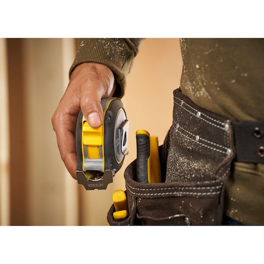 Photo of STANLEY FMHT38232-0 tape measure held by worker next to tool belt with yellow tools.