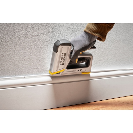 Stanley FMHT70100-0 cable tacker in use along baseboard, shown held by a gloved hand against wall.