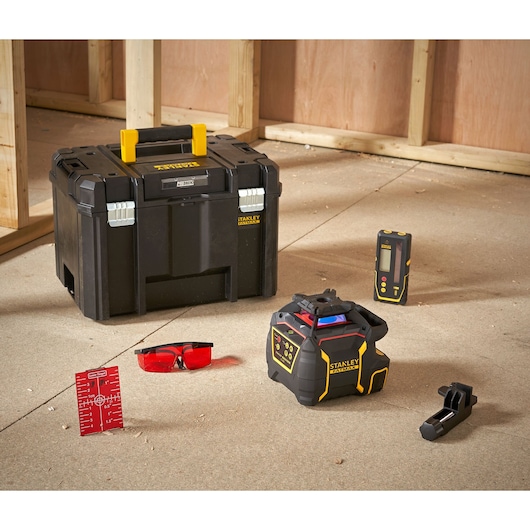 STANLEY tool set for professional photography and measuring, including a black toolbox with yellow handle, a laser level device, a laser detector, a clamp, red tinted safety glasses, and a red target sheet, arranged on a construction site floor with wooden framing in the background.