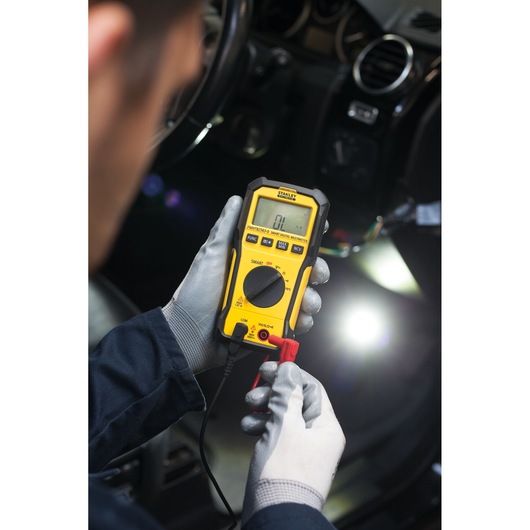 Photo of STANLEY FMHT82563-0 digital multimeter used for car diagnostics, held by gloved hands.