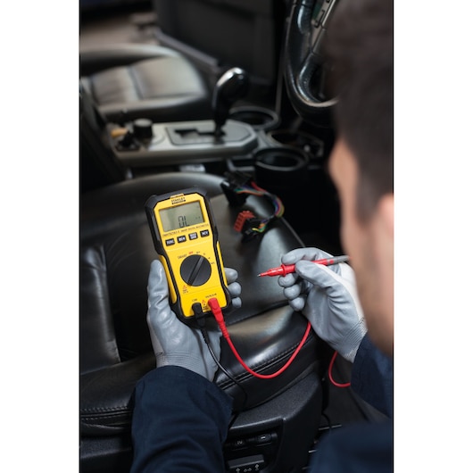 Photo of STANLEY FMHT82563-0 digital multimeter in use inside a car, with gloved hands and test leads.