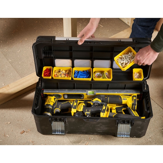 Open STANLEY tool box showing organized compartments with screws, wall plugs, tape, and several yellow and black STANLEY power tools inside. A person's hand is lifting a tray of screws.