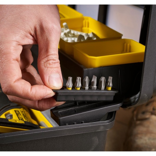 Close-up of a hand picking up a set of screwdriver bits from a black and yellow organizer box, with several compartments and tools visible.