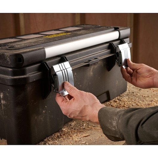 Hands opening the metal latches on a dusty black STANLEY toolbox with a white handle, placed on a workbench covered in wood shavings.