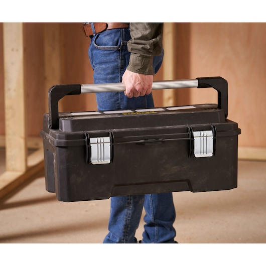 A person carrying a black Stanley toolbox with a large silver handle and secure metal latches, inside a construction environment.