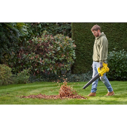 STANLEY SFMCBL714B-XJ leaf blower in use on grass, blowing a pile of dry leaves in a garden setting.