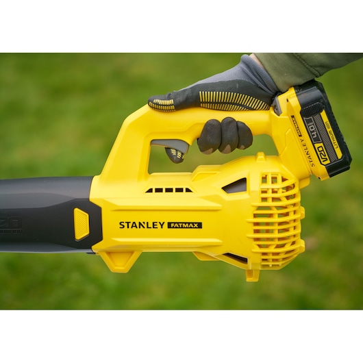 Photo of STANLEY SFMCBL714B-XJ cordless leaf blower held by gloved hand, yellow and black design outdoors.