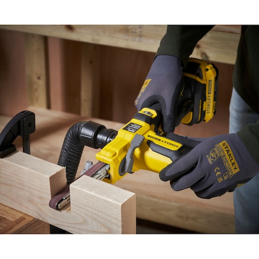 Photo of STANLEY SFMCE210B cordless belt sander in use on a wood surface, worker wearing gloves.