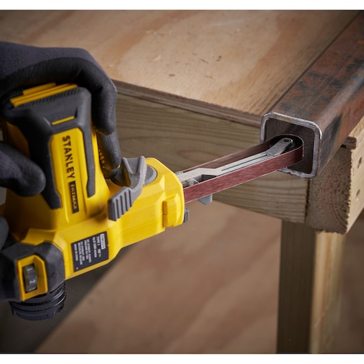 STANLEY SFMCE210B cordless belt file sander smoothing inside metal pipe, side angle, held by gloved hand.