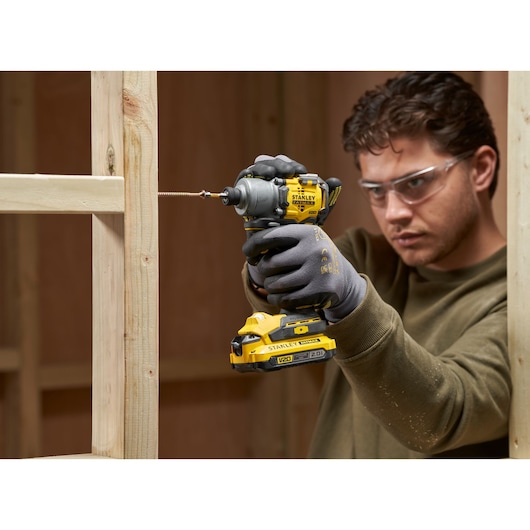 Photo of STANLEY SFMCF830B impact driver in use, driving screw into wooden frame, user wearing gloves.
