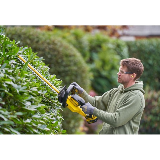 Photo of STANLEY SFMCHT655B hedge trimmer in use, yellow and black, trimming green bushes outdoors.