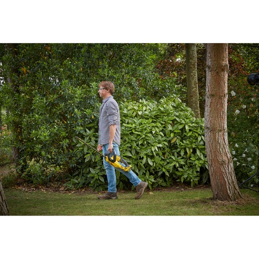 Photo of STANLEY SFMCHT855B hedge trimmer held by person walking outdoors near greenery.