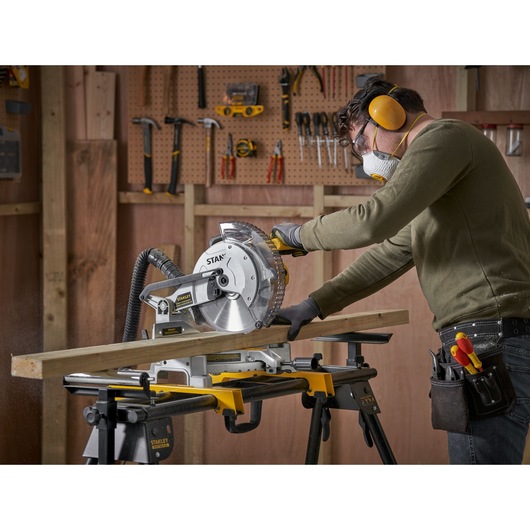 Photo of a person using the STANLEY SM1650 miter saw to cut wood, in a workshop with tools on a pegboard wall.