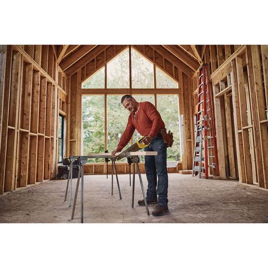 STANLEY STHT20348 hand saw used to cut wood plank on sawhorses inside a wooden house frame under construction.