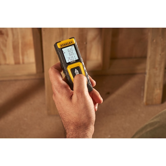 Photo of STANLEY STHT77100-0 laser distance measurer held in hand, display screen visible in construction setting.