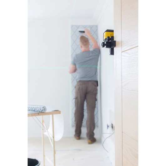 Photo of STANLEY STHT77649-1 laser level mounted on wall in home renovation scene, blurred person hanging wallpaper.