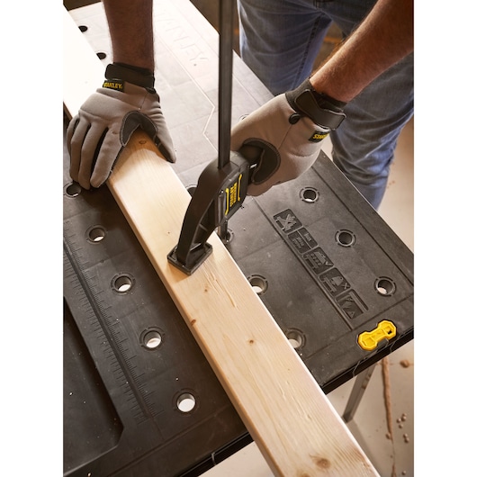Photo of STANLEY STST83492-1 workbench with gloved hands clamping wood plank on black surface.